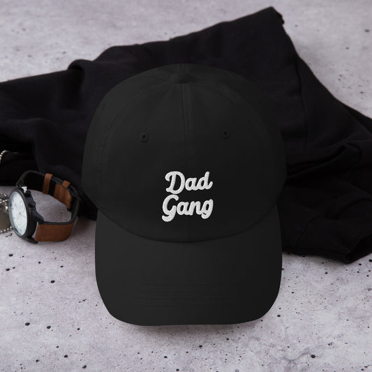 Black cap with Dad Gang embroidered in thick cursive font laid next to a hoodie and leather-band watch 